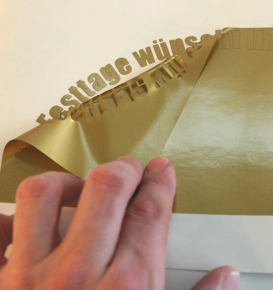 Hand zieht überschüssige Goldfolie ab, der geplottete Schriftzug Festtage wünscht bleibt beim Entgittern auf dem weißen Trägerpapier