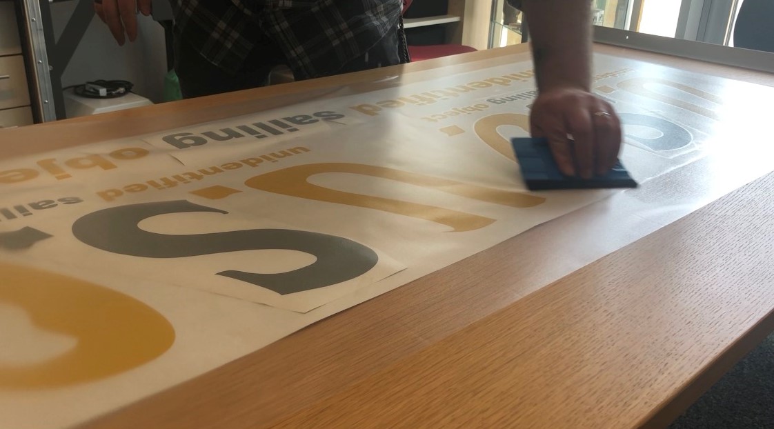 Hand streicht mit blauem Rakel Übertragungsfolie auf geplottete Schrift U.S.O. in Gold und Grau, zwei Folienfarben gemeinsam präpariert