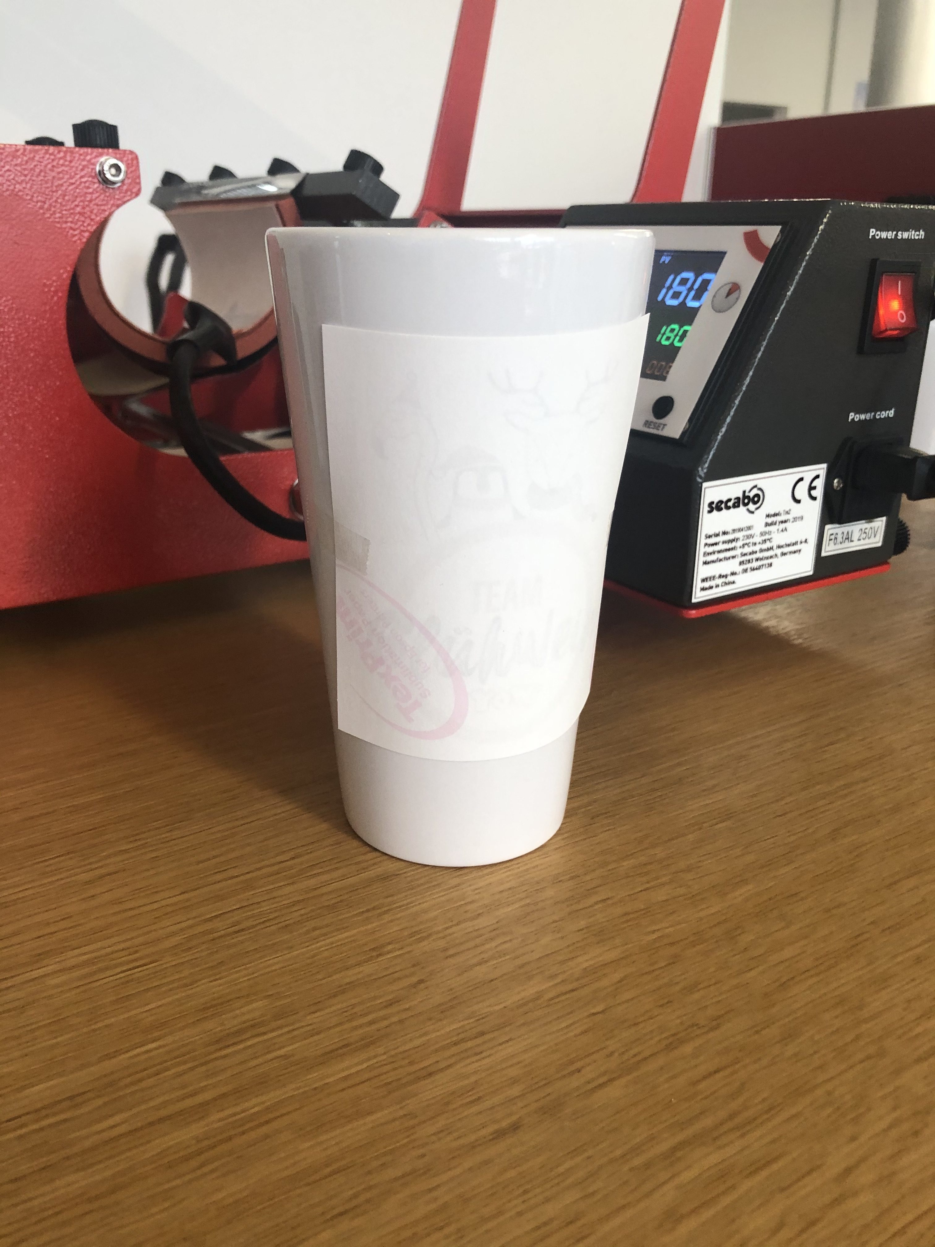 Weiße Tasse mit per Thermoband fixiertem Sublimationspapier vor roter Secabo-Tassenpresse, Display zeigt 180 °C