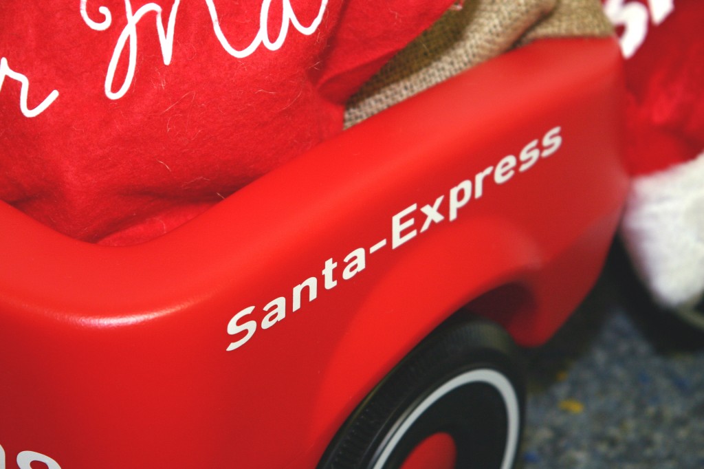 Roter Bobby-Car-Anhänger mit geplottetem weißem Folienschriftzug Santa-Express, beladen mit rotem Filzsack und Jutesack als Nikolausdeko