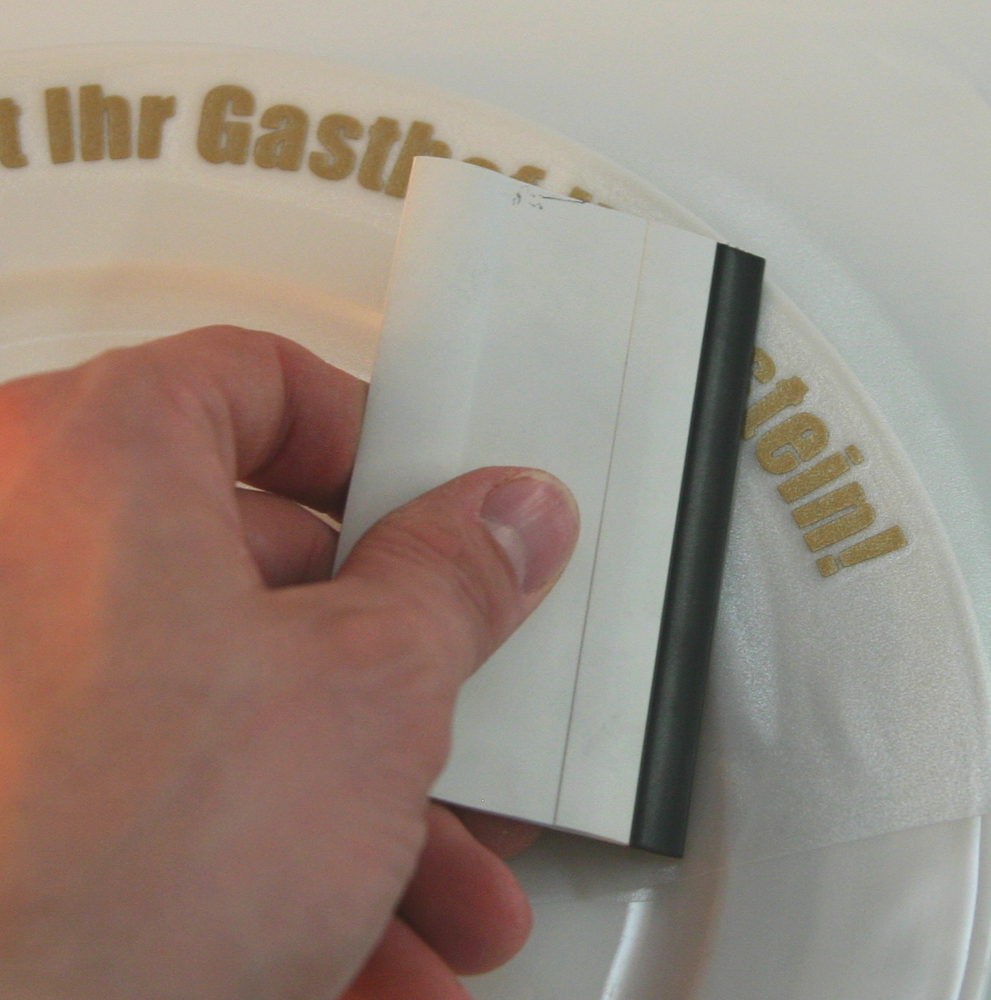 Hand drückt goldenen Folienschriftzug mit einem Rakel durch die Übertragungsfolie auf den Rand eines weißen Tellers fest