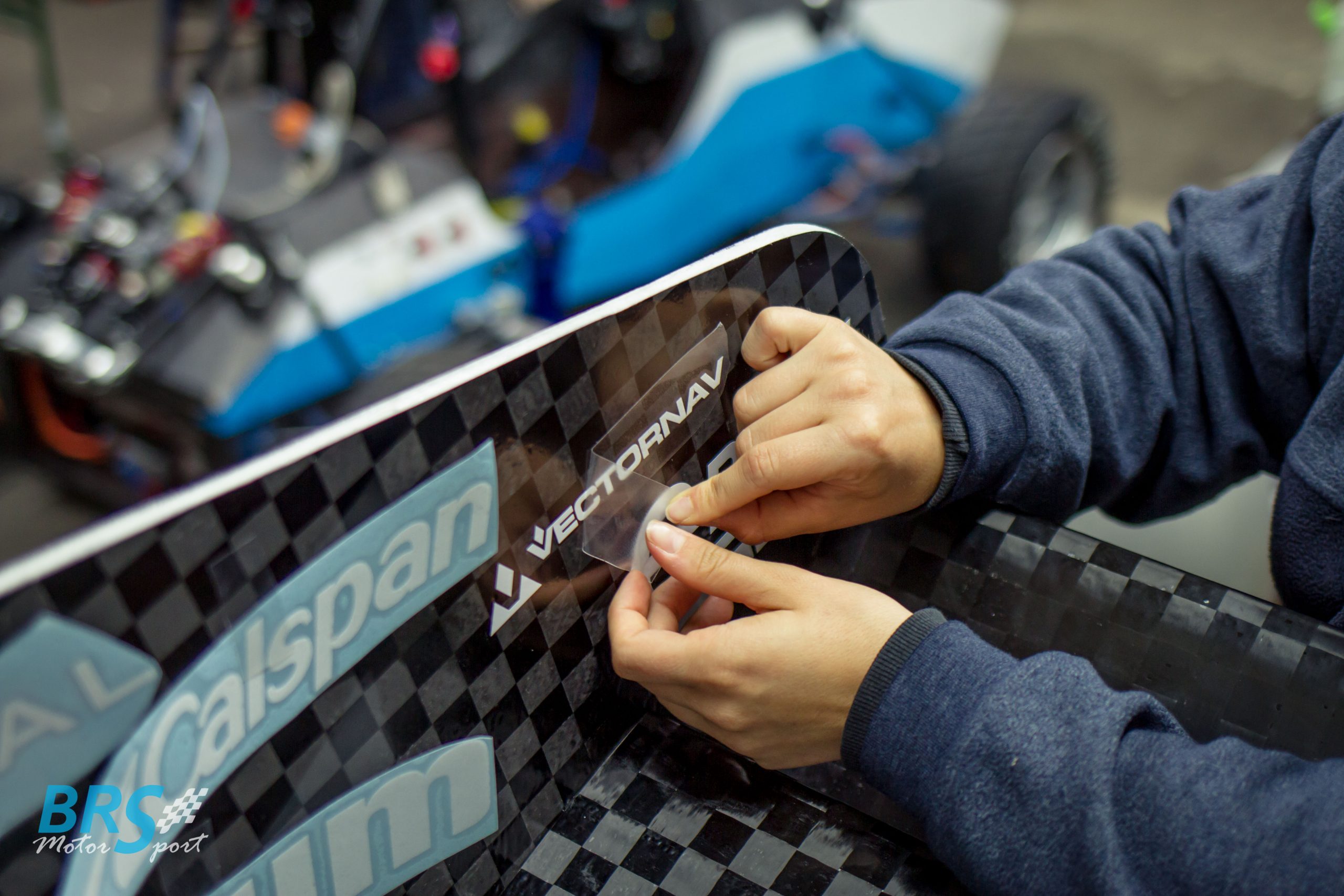 Hände ziehen Transferfolie von geplottetem VectorNav-Schriftzug auf Carbonteil eines BRS-Motorsport-Rennwagens ab, daneben Calspan-Aufkleber