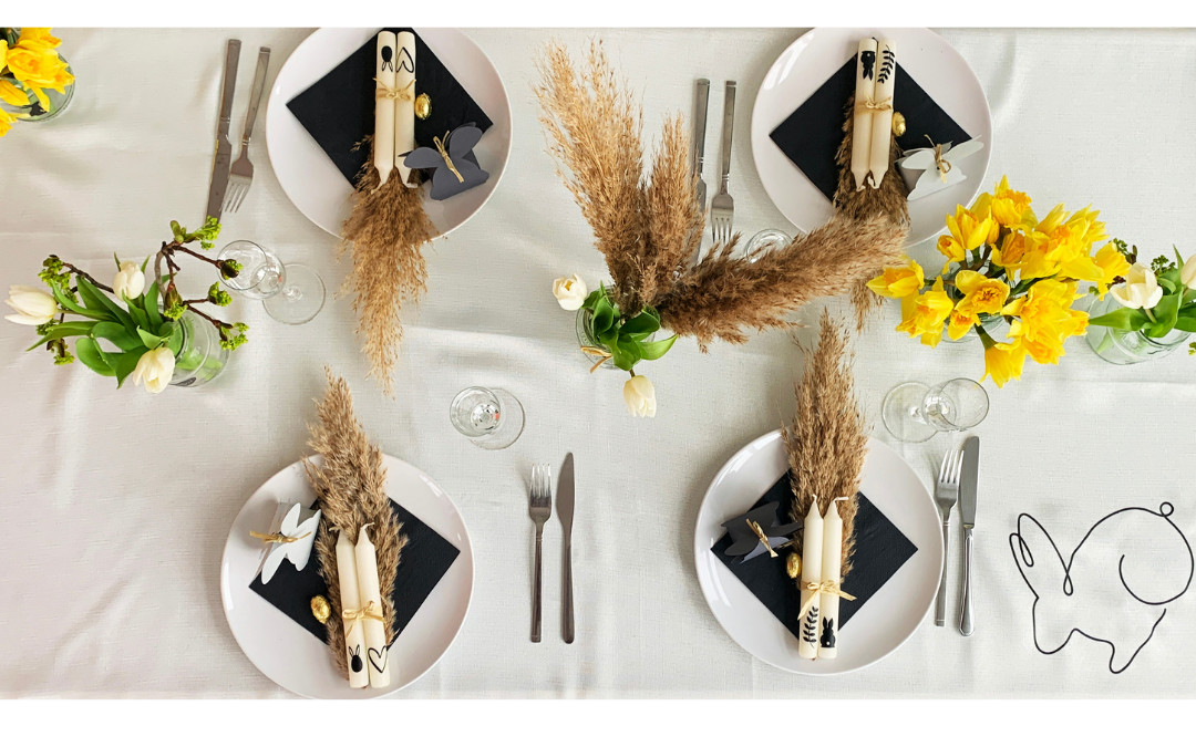 Ostertafel von oben: weiße Tischdecke mit dezentem Hasen-Linienprint, Teller mit schwarzen Servietten, bemalten Kerzen, Pampasgras und Narzissen