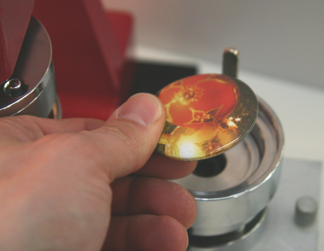 Hand hält fertig gepressten Button mit roter Weihnachtskugel-Motiv vor der silbernen Presseinheit einer roten Buttonmaschine