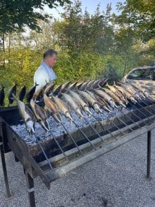 Steckerlfische auf Spießen über glühender Holzkohle am Grill, dahinter ein Mann im hellen Hemd, bei der Nepata Wiesn 2021 im Freien