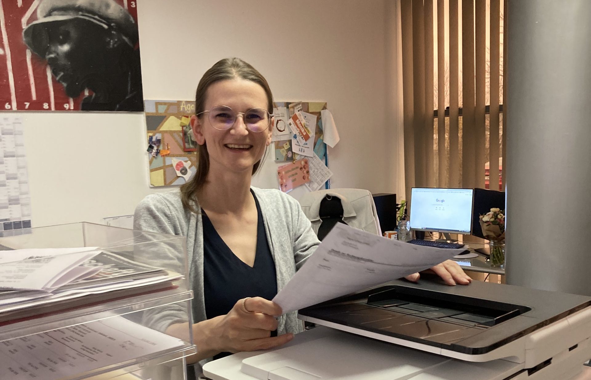 Moni lächelt mit Unterlagen in der Hand am Drucker in ihrem Büro bei Nepata, hinter ihr eine Pinnwand und Blumen auf dem Schreibtisch