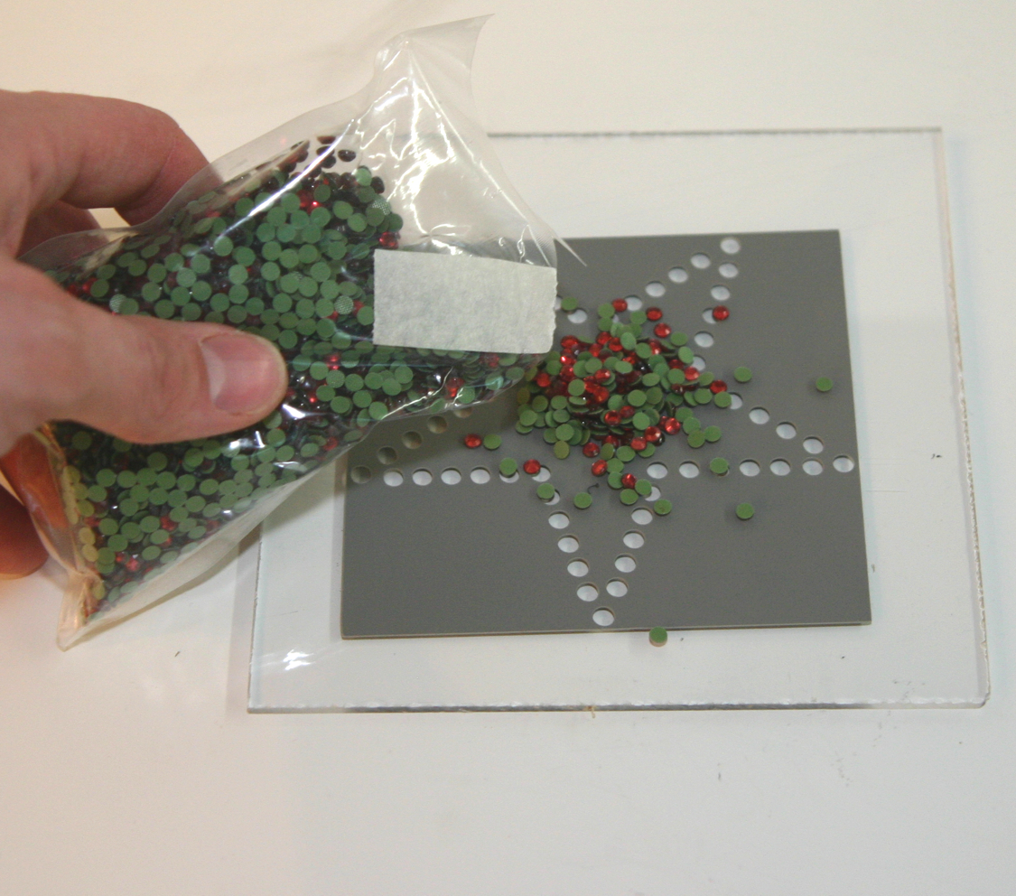 Hand schüttet Strass-Steine aus einem Beutel auf eine graue Schablonenfolie mit Stern aus Löchern, aufgeklebt auf Acrylglas