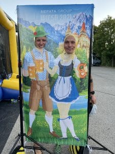 Zwei Gäste stecken ihre Köpfe durch eine Fotowand der Nepata Group Wiesn 2021 mit gemaltem Trachtenpaar, Bierkrügen und Brezn