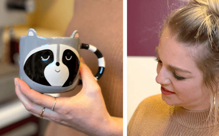 Hand hält eine graue Tasse mit Waschbärgesicht, daneben lächelt Sabrina mit blonden Haaren im senfgelben Strickpullover nach unten