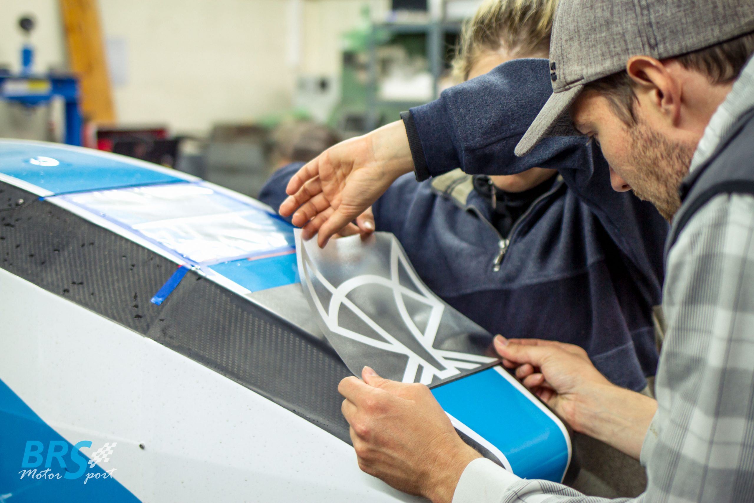 Zwei Studierende von BRS Motorsport bringen in der Werkstatt einen weißen Folienschnitt per Transferfolie auf die Carbon-Karosserie ihres Rennwagens auf