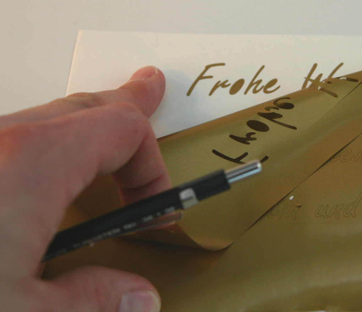 Hand mit Stift zieht goldene Plotterfolie von weißer Karte ab, zurück bleibt der goldene Schriftzug Frohe Weihnachten