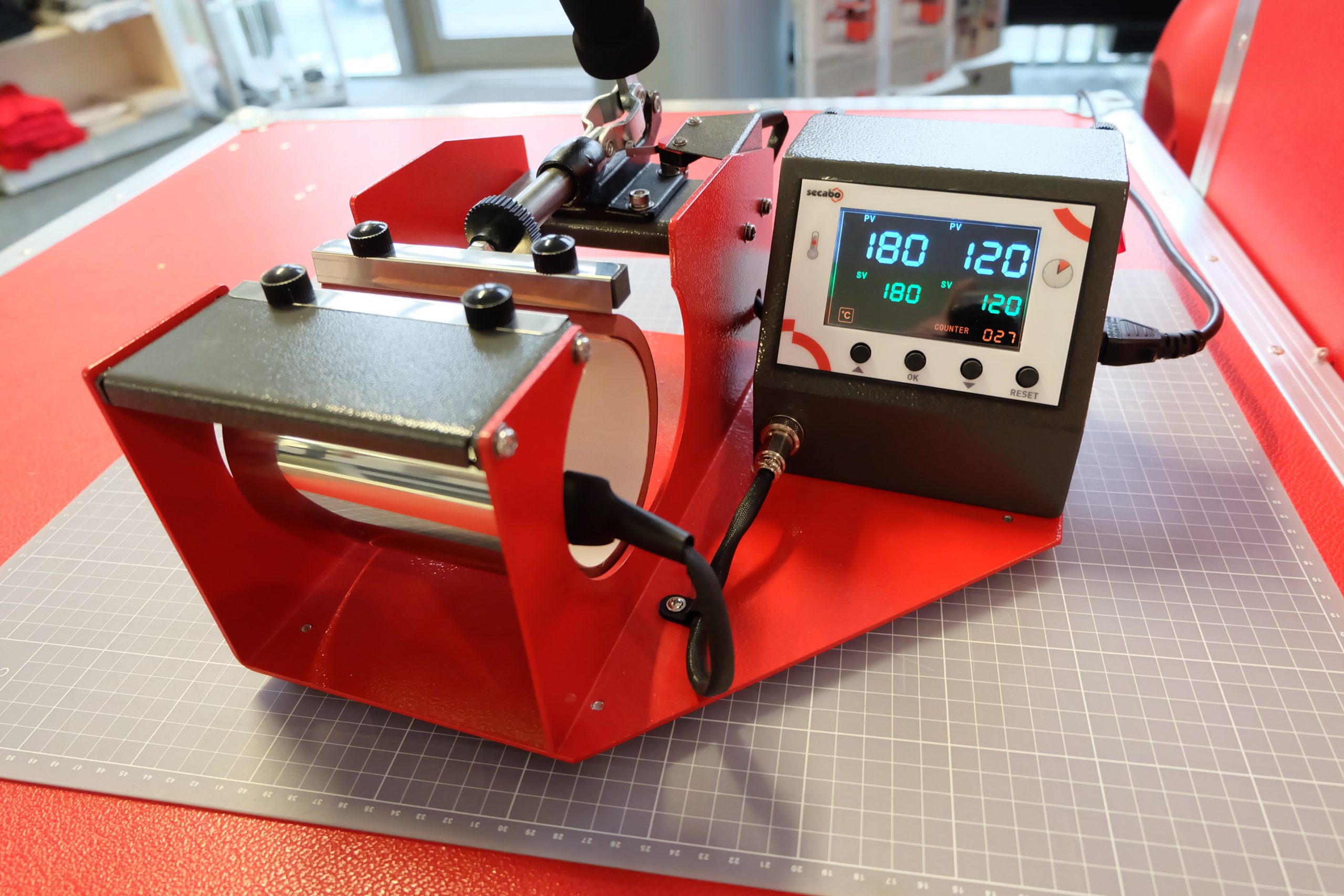 Rote Secabo TM1 Tassenpresse mit eingelegtem Edelstahlbecher, Display zeigt 180 °C und 120 Sekunden als Pressparameter
