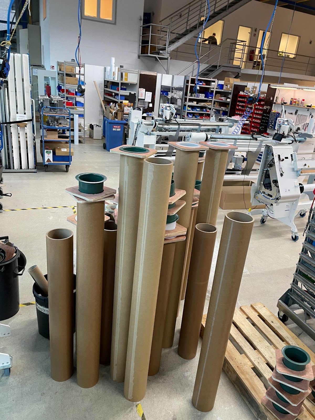 Aufrecht stehende 6-Zoll-Papphülsen, teils mit grünen Kunststoffeinsätzen auf Holzplatten, in der NEPATA-Werkstatt vor Umwickelmaschinen