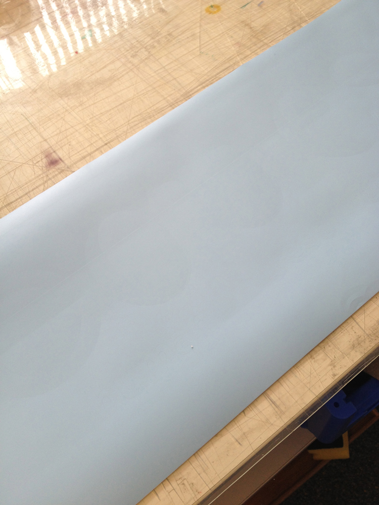 Hellblaue Plotterfolie auf zerkratztem Schneidetisch, darin schwach sichtbare Schnittkonturen aller Motivteile – erste Farbe vollflächig geplottet