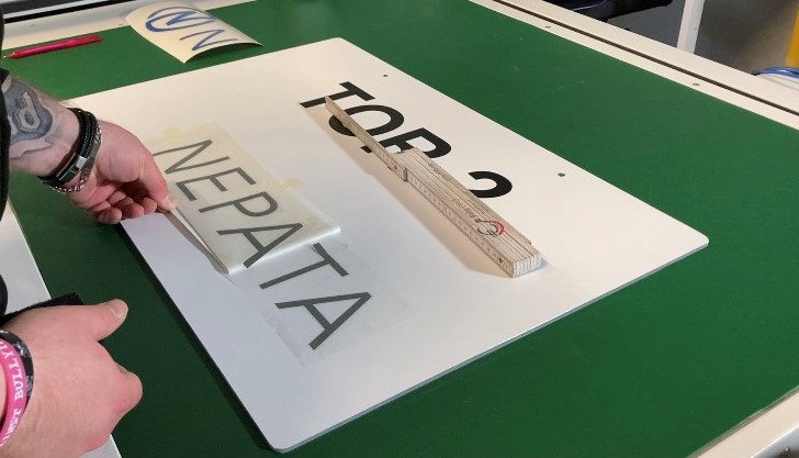 Hände kleben Schriftzug mit Übertragungsfolie auf weißes Schild mit Aufschrift TOR 2, Zollstock als Ausrichthilfe auf grünem Arbeitstisch