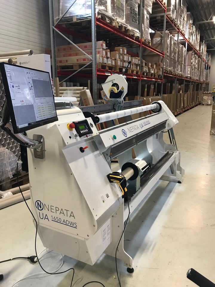 Machine d'emballage sous film NEPATA UA 1650 ADWS, équipée d'un écran pour le logiciel de commande et d'un scanner portable, dans un entrepôt à rayonnages en hauteur