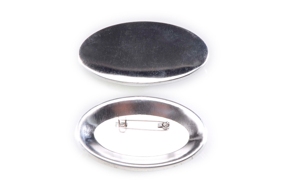 Zwei unbedruckte ovale Metall-Buttons im Format 45 x 69 mm: oben die glatte Vorderseite, unten die Rückseite mit Sicherheitsnadel