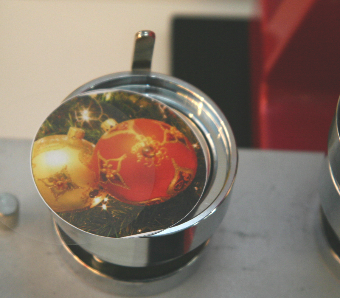 Ausgestanztes Motiv mit rot-goldenen Christbaumkugeln und Klarsichtfolie in der Metallaufnahme einer Buttonmaschine, bereit zum Pressen