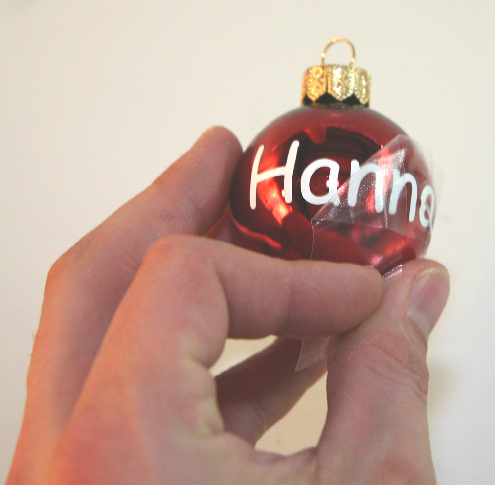 Rote Christbaumkugel mit weißem Schriftzug Hanna aus Plotterfolie, von der eine Hand die transparente Übertragungsfolie abzieht