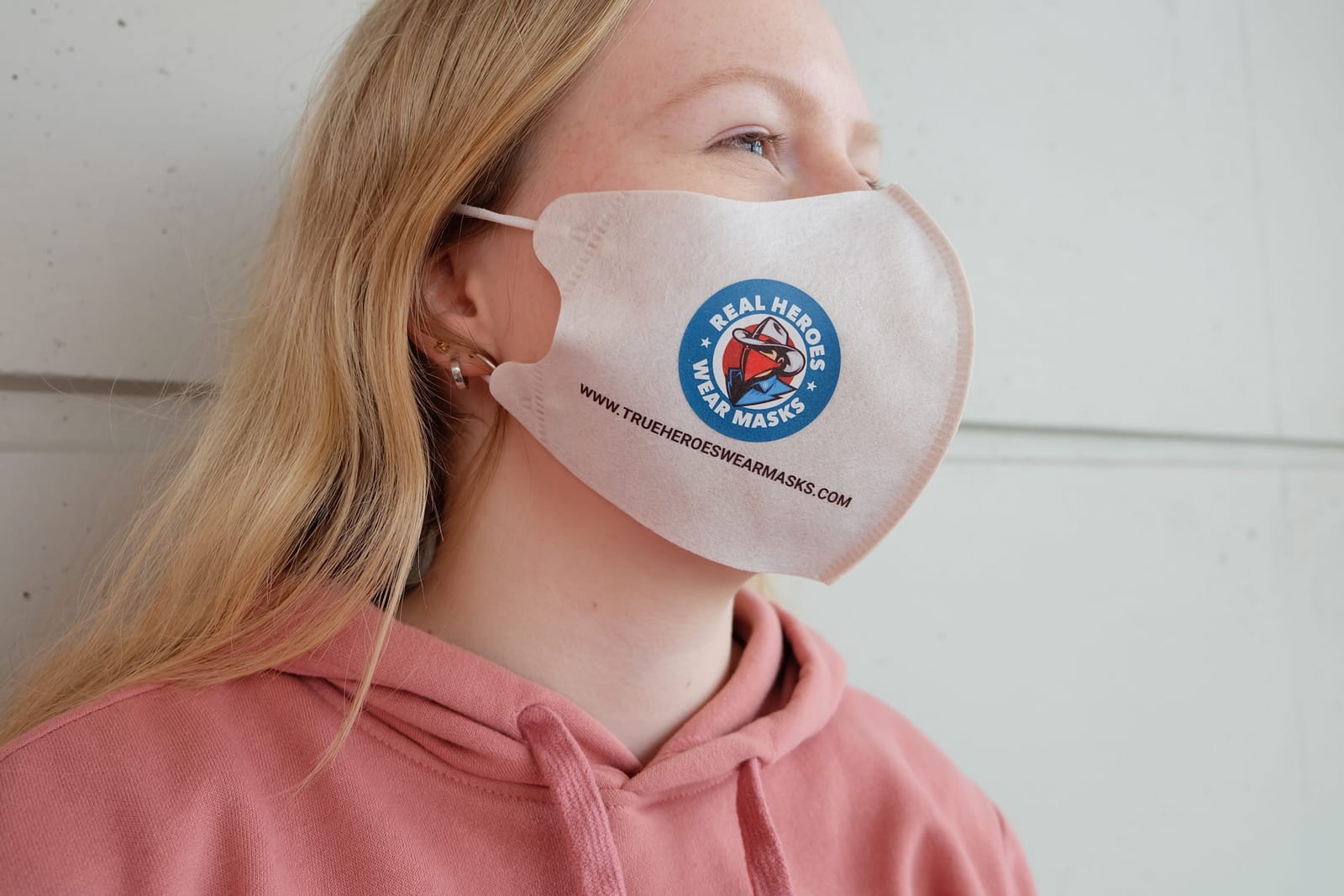 A woman is wearing a white non-woven mask printed with the ‘Real Heroes Wear Masks’ and ‘www.trueheroeswearmasks.com’ logos – an example of masks with a message