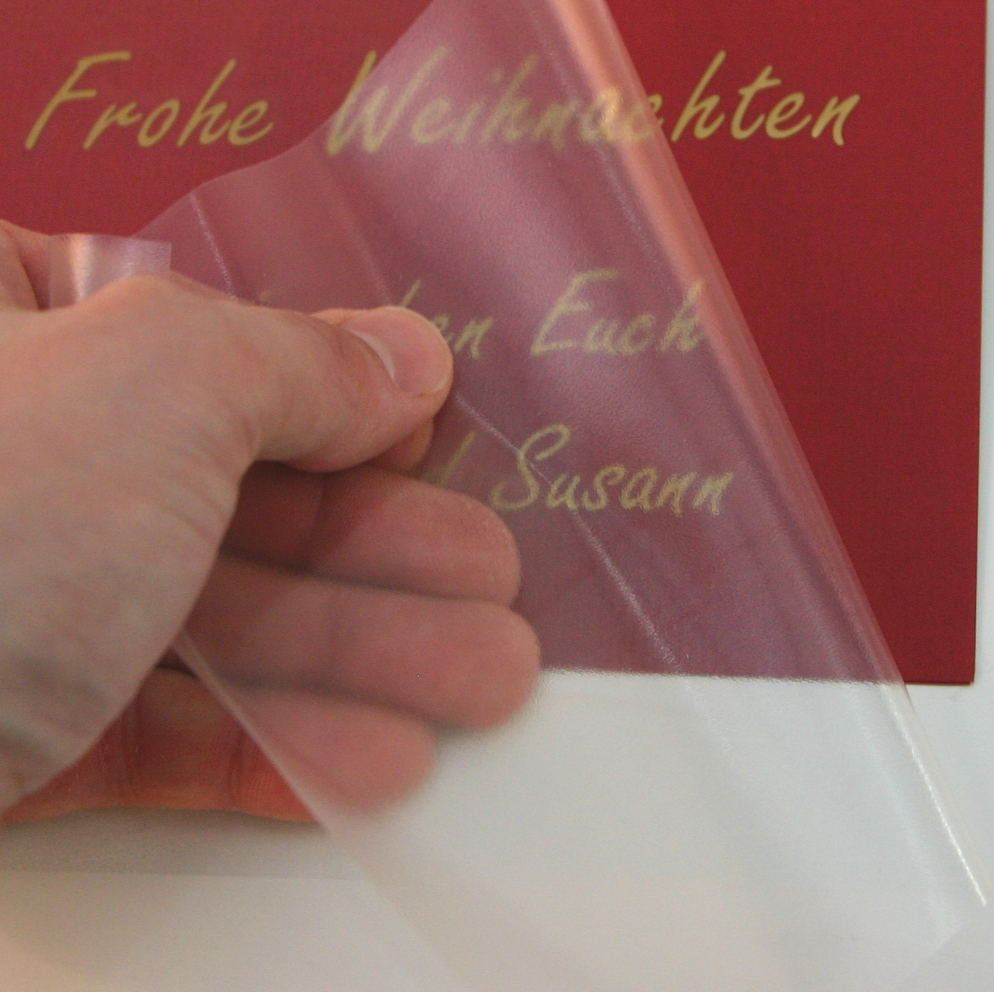 Hand zieht transparente Applikationsfolie von roter Karte ab, darauf bleibt der goldene geplottete Schriftzug Frohe Weihnachten haften
