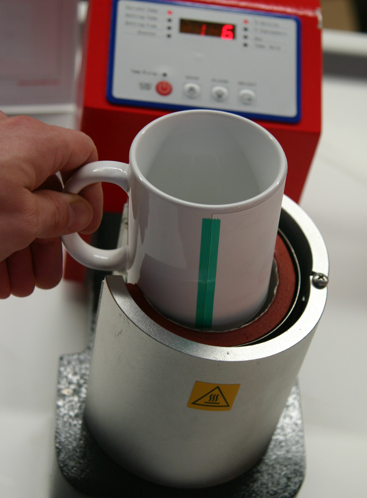 Hand setzt weiße Tasse mit grünem Klebeband in die zylindrische Transferpresse zum Sublimieren, daneben rotes Bedienteil mit Digitalanzeige