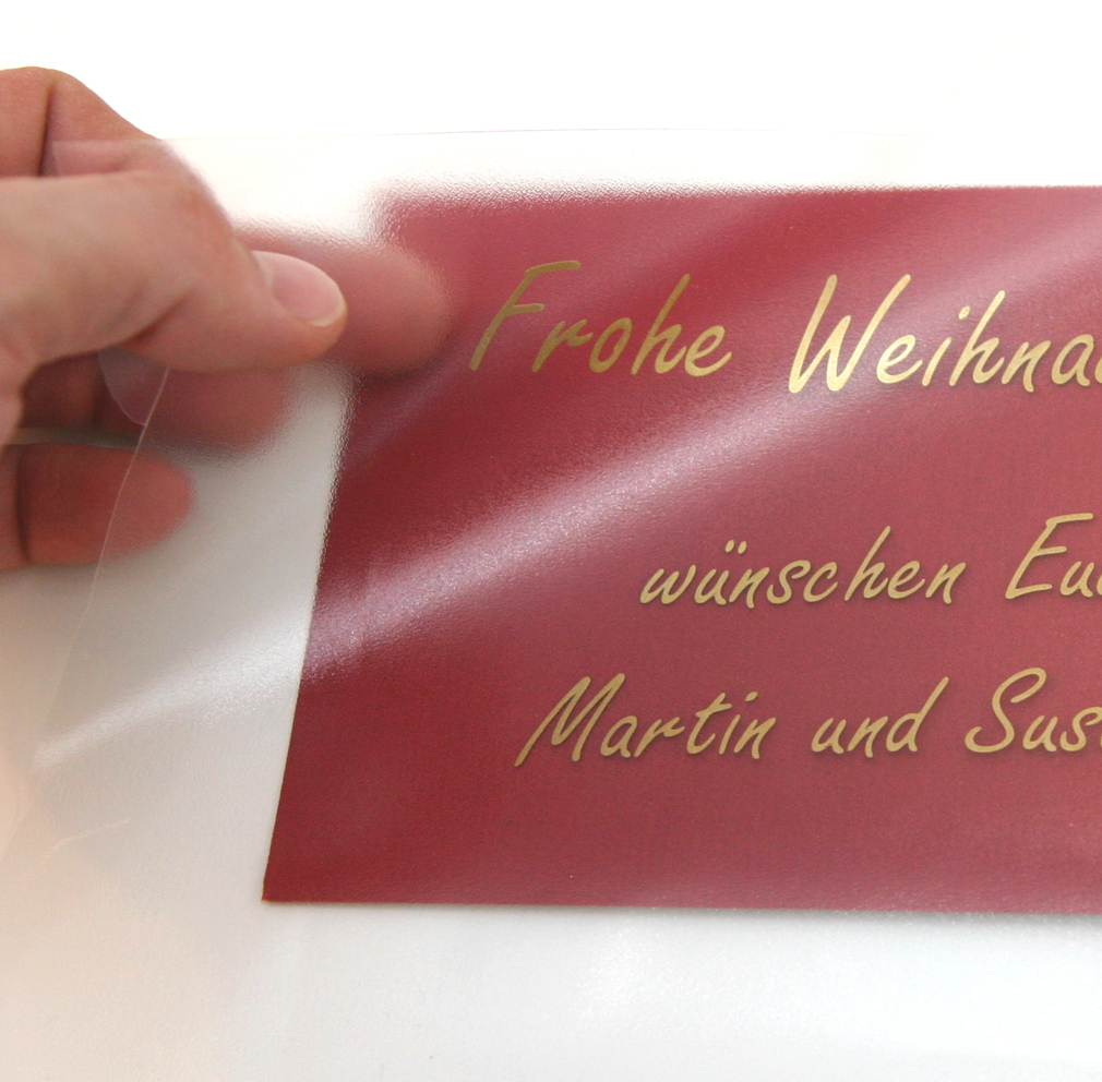 Hand zieht transparente Applikationsfolie von roter Karte mit goldenem, geplottetem Schriftzug Frohe Weihnachten … Martin und Sus…