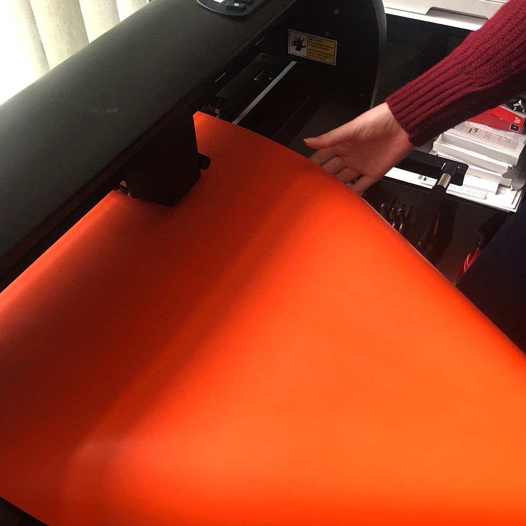 Hand legt orange Flexfolie in einen schwarzen Schneideplotter ein, um das Trick-or-Treat-Motiv für die Halloween-Tüte zu plotten