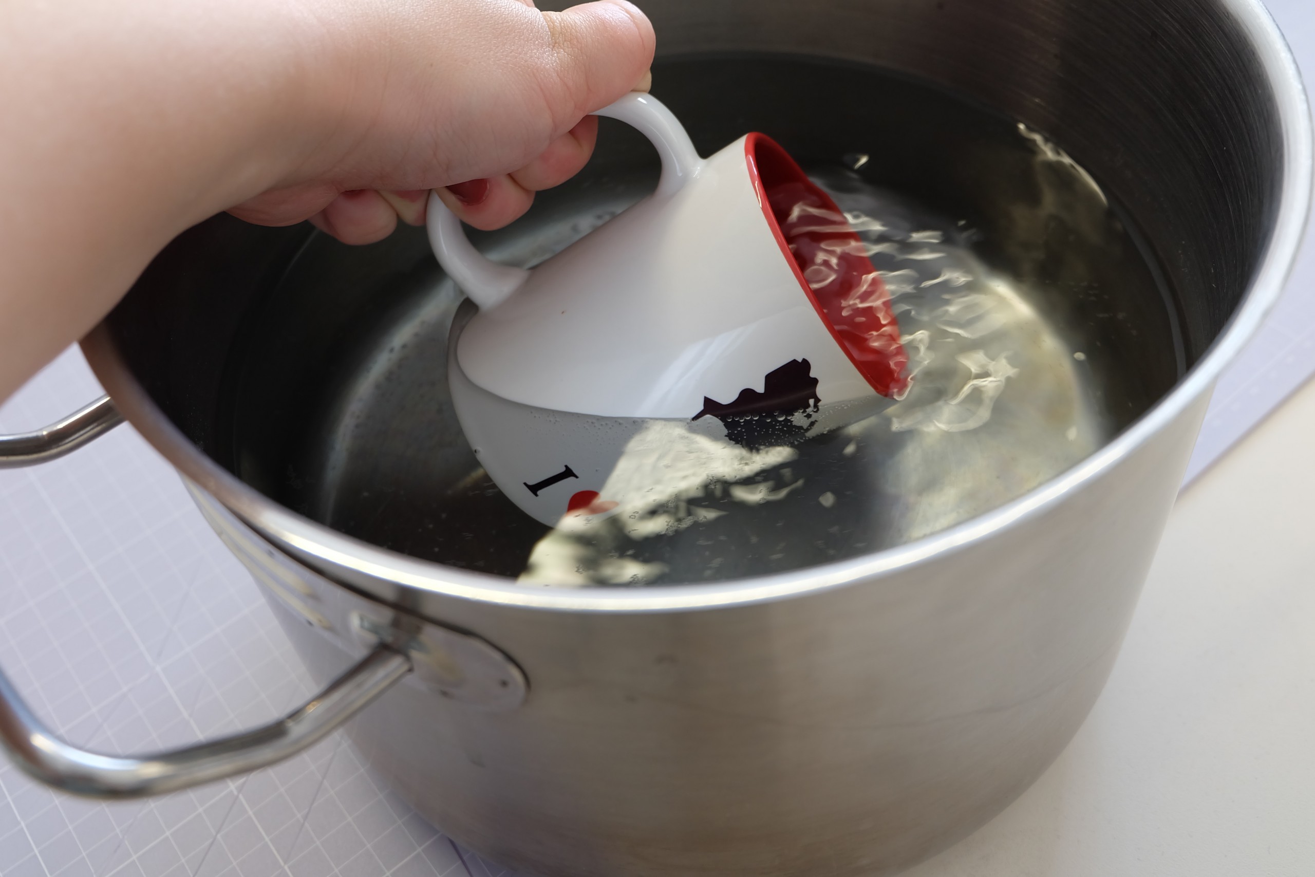 Hand taucht bedruckte weiße Tasse mit rotem Innenrand in Edelstahltopf mit heißem Wasser, um sie nach dem Sublimationsdruck abzuschrecken