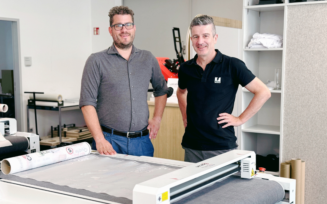 Zwei Männer stehen hinter dem Secabo DTF MultiCut Pro, einem Flachbett-Schneideplotter mit bedruckter Folienrolle, in einem Showroom