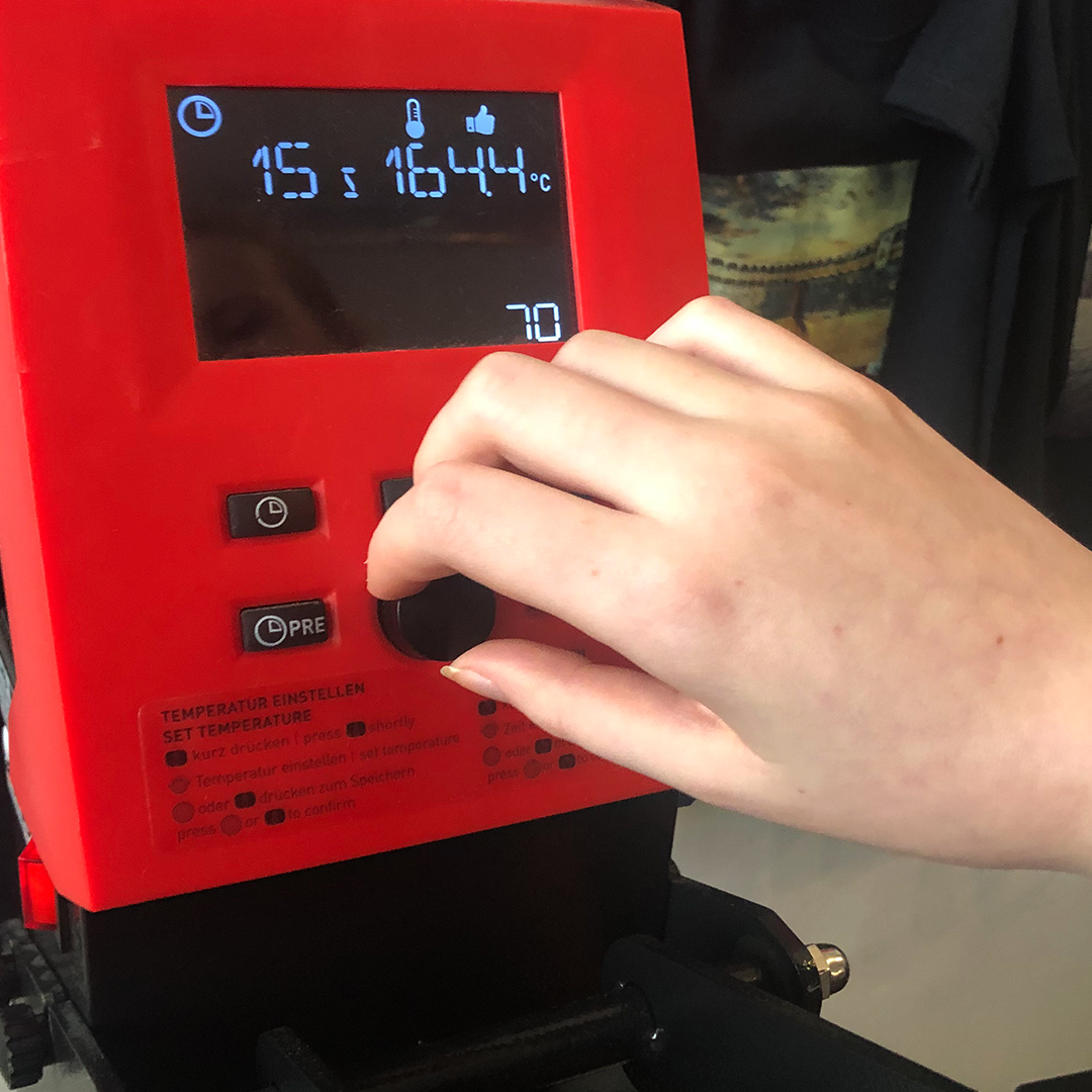 Hand stellt am roten Bedienfeld einer Transferpresse Anpressdauer und Temperatur ein, das Display zeigt 15 s und 164,4 °C