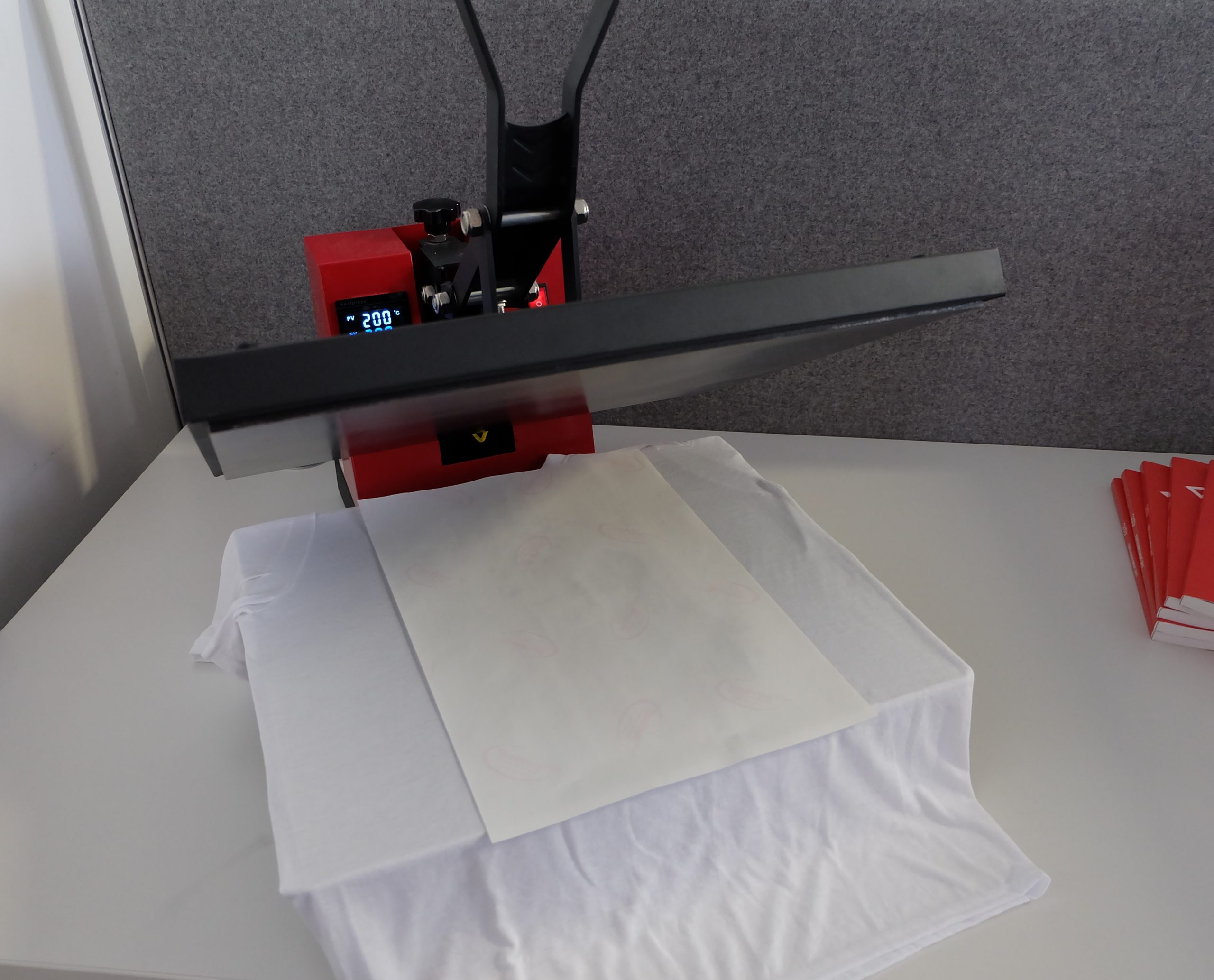 Geöffnete rote Transferpresse mit Anzeige 200 °C, darunter weißes T-Shirt mit aufgelegtem bedrucktem Sublimationspapier