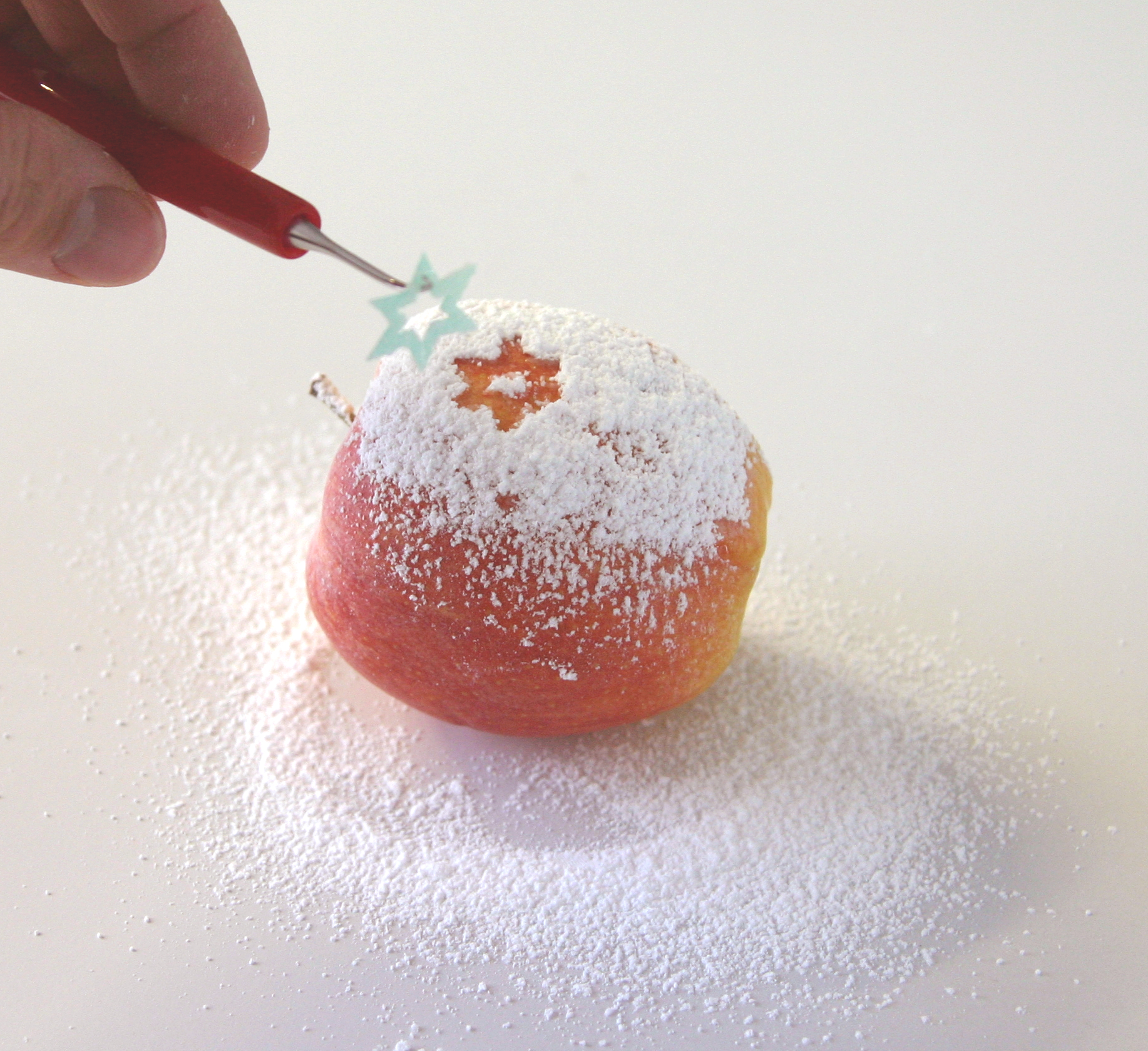 Mit Puderzucker bestäubter Apfel, ein Entgitterhaken zieht einen grünen Sternaufkleber ab und legt ein rotes Sternmotiv frei