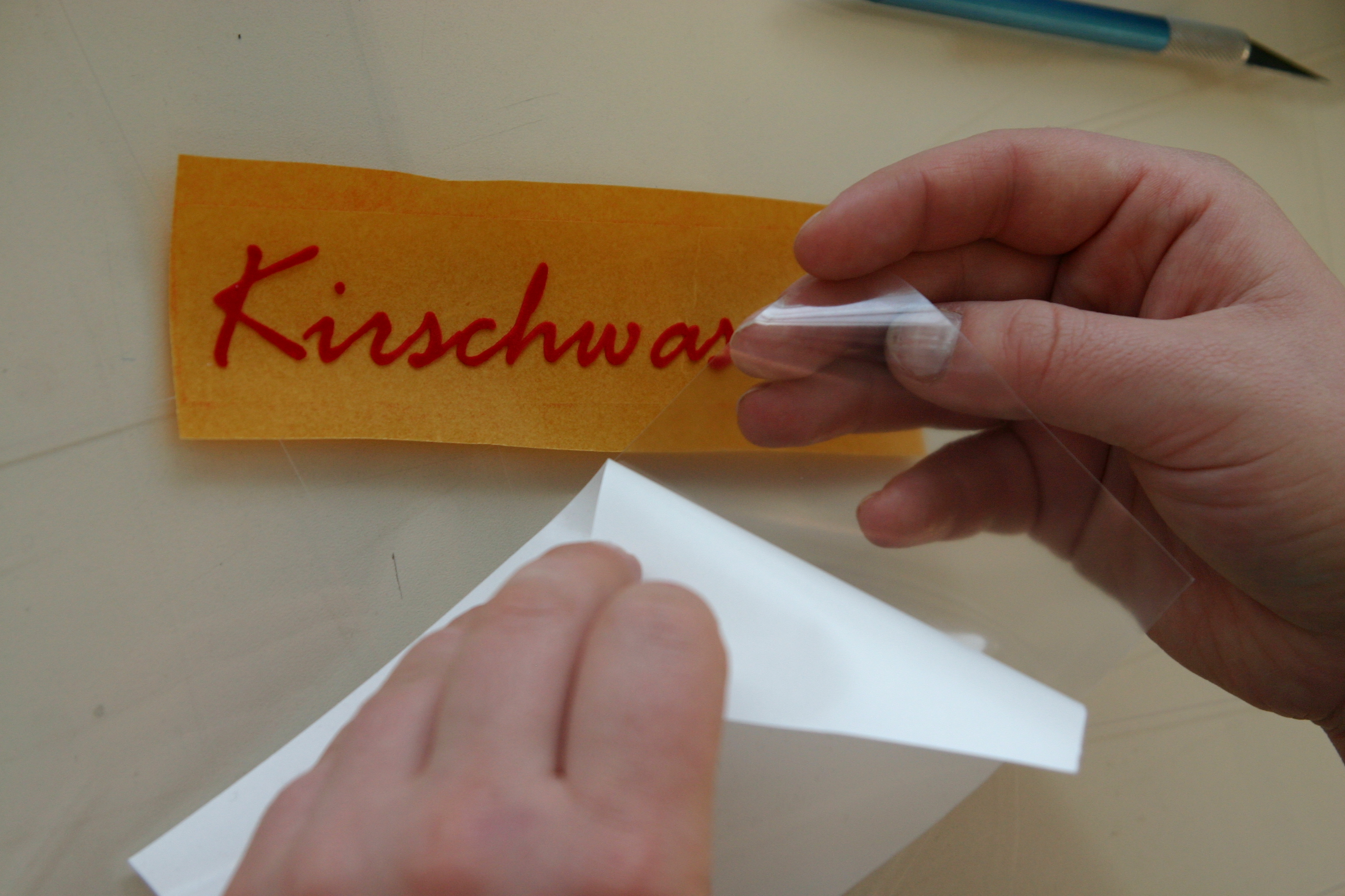 Hände lösen das Trägerpapier vom roten Flockfolien-Schriftzug Kirschwasser mit Übertragungsfolie, daneben ein Cutter
