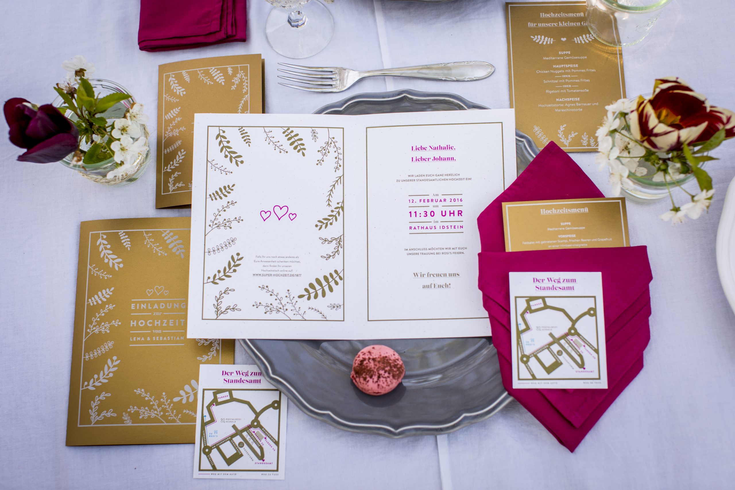 Hochzeitspapeterie auf gedecktem Tisch: goldene Einladungen und Menükarten mit weiß gedrucktem Blätterdekor, dazu Anfahrtskarten und pinke Servietten