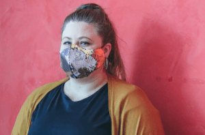 Frau mit gemusterter Stoffmaske, dunklem Shirt und senfgelber Strickjacke vor roter Wand, Symbol für den Alltag mit Maskenpflicht ab Mai 2020