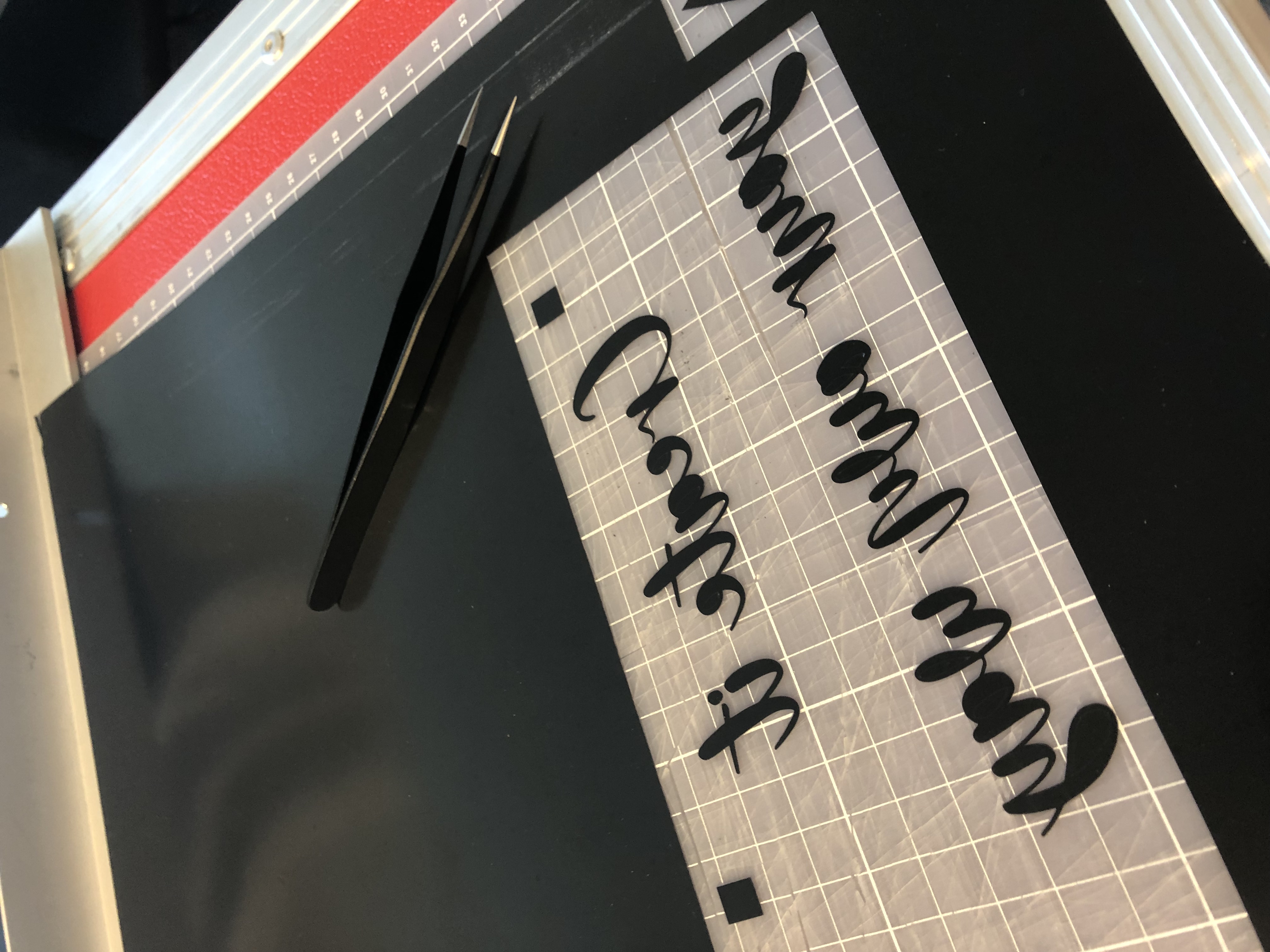 Entgitterte schwarze Flexfolie mit gespiegeltem Schriftzug Create it your own way auf transparenter Trägerfolie, daneben eine Pinzette
