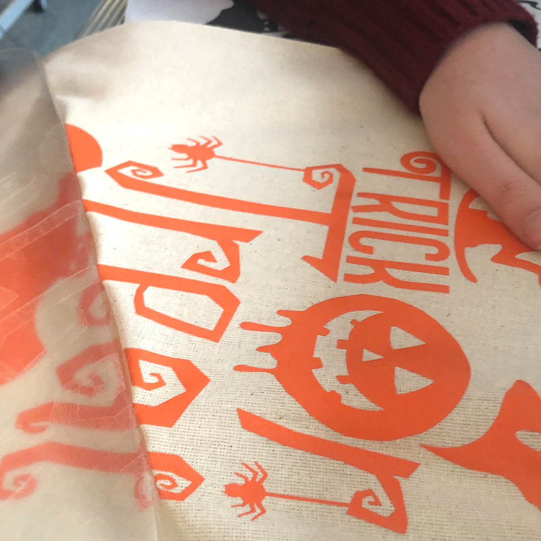 Trägerfolie wird von orangefarbenem Flexfolien-Motiv mit Kürbis und Trick-or-Treat-Schriftzug auf Baumwolltasche abgezogen