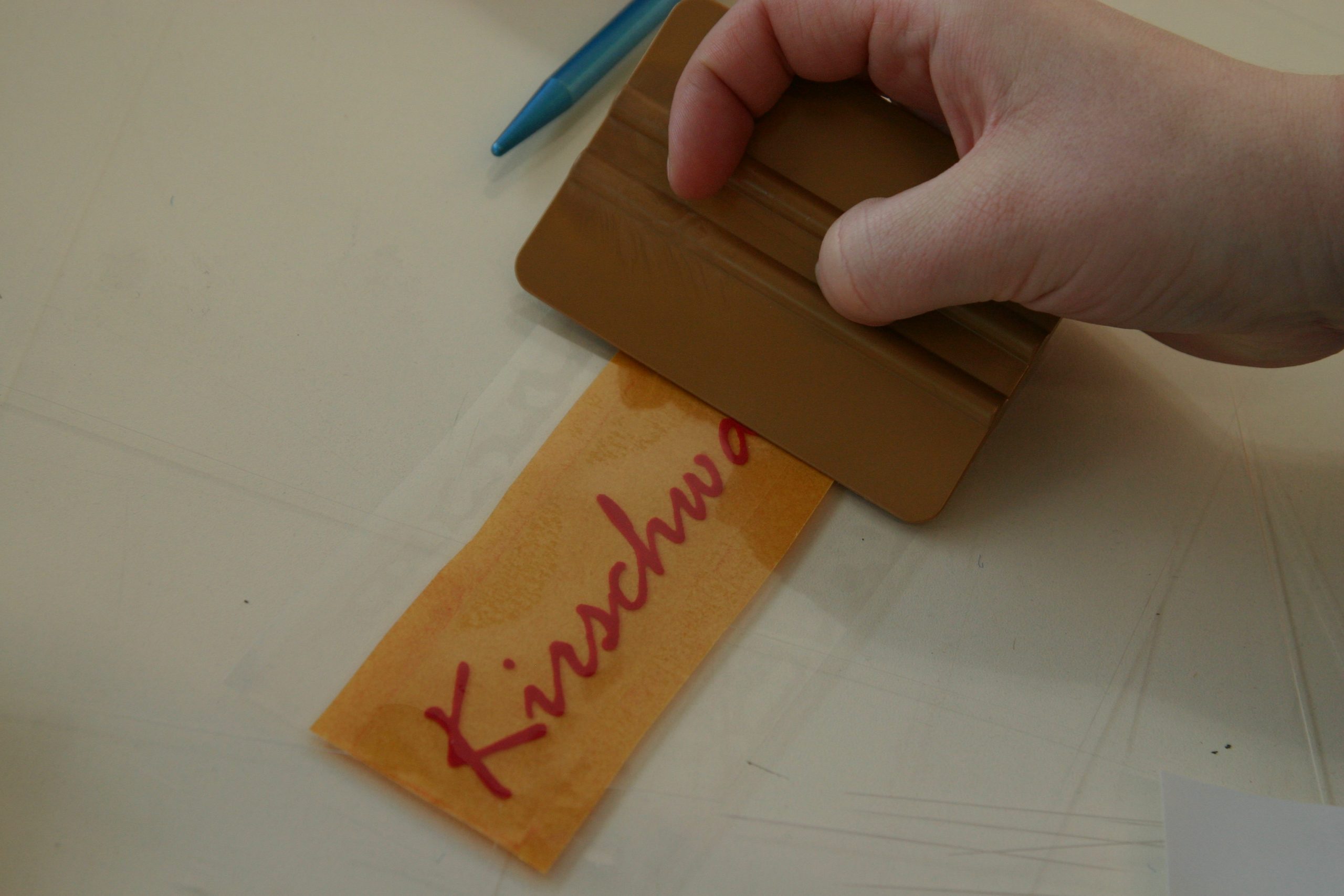 Hand streicht mit einer Rakel orange Übertragungsfolie auf den entgitterten roten Flockfolien-Schriftzug Kirschwa…, daneben Entgitternadel