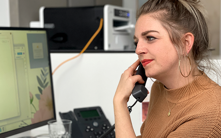 Sabrina im braunen Strickpullover telefoniert an ihrem Arbeitsplatz und blickt auf einen Monitor mit geöffnetem Grafikentwurf