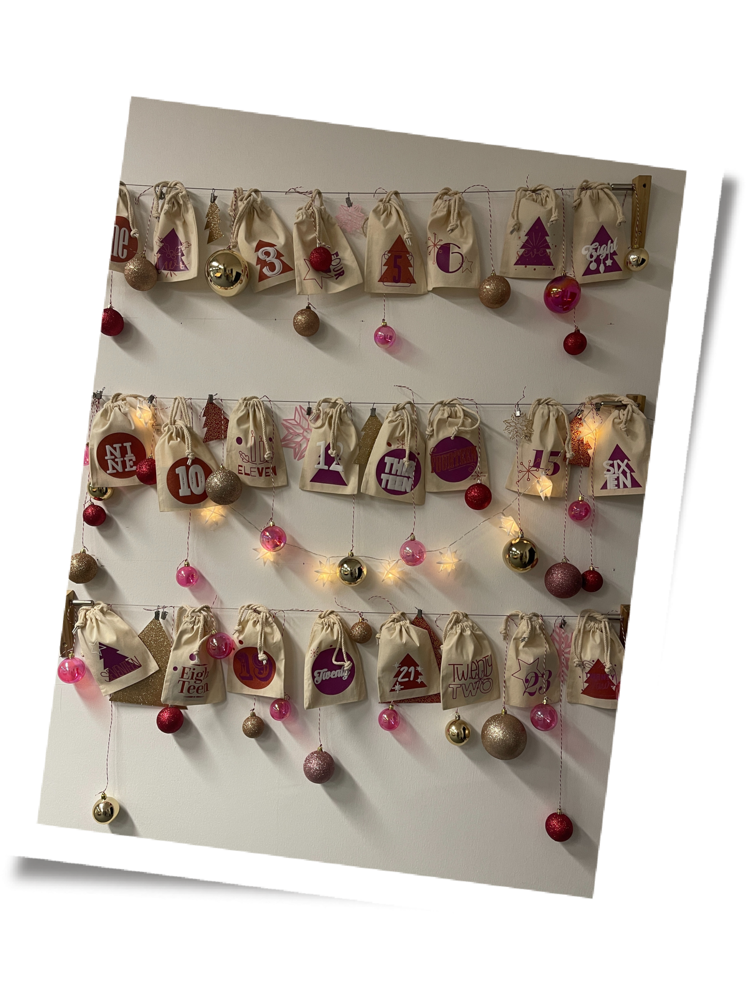 DIY-Adventskalender: 24 Stoffsäckchen mit pinken und roten Zahlenmotiven an drei Drahtseilen, dazu Christbaumkugeln und Sternenlichterkette