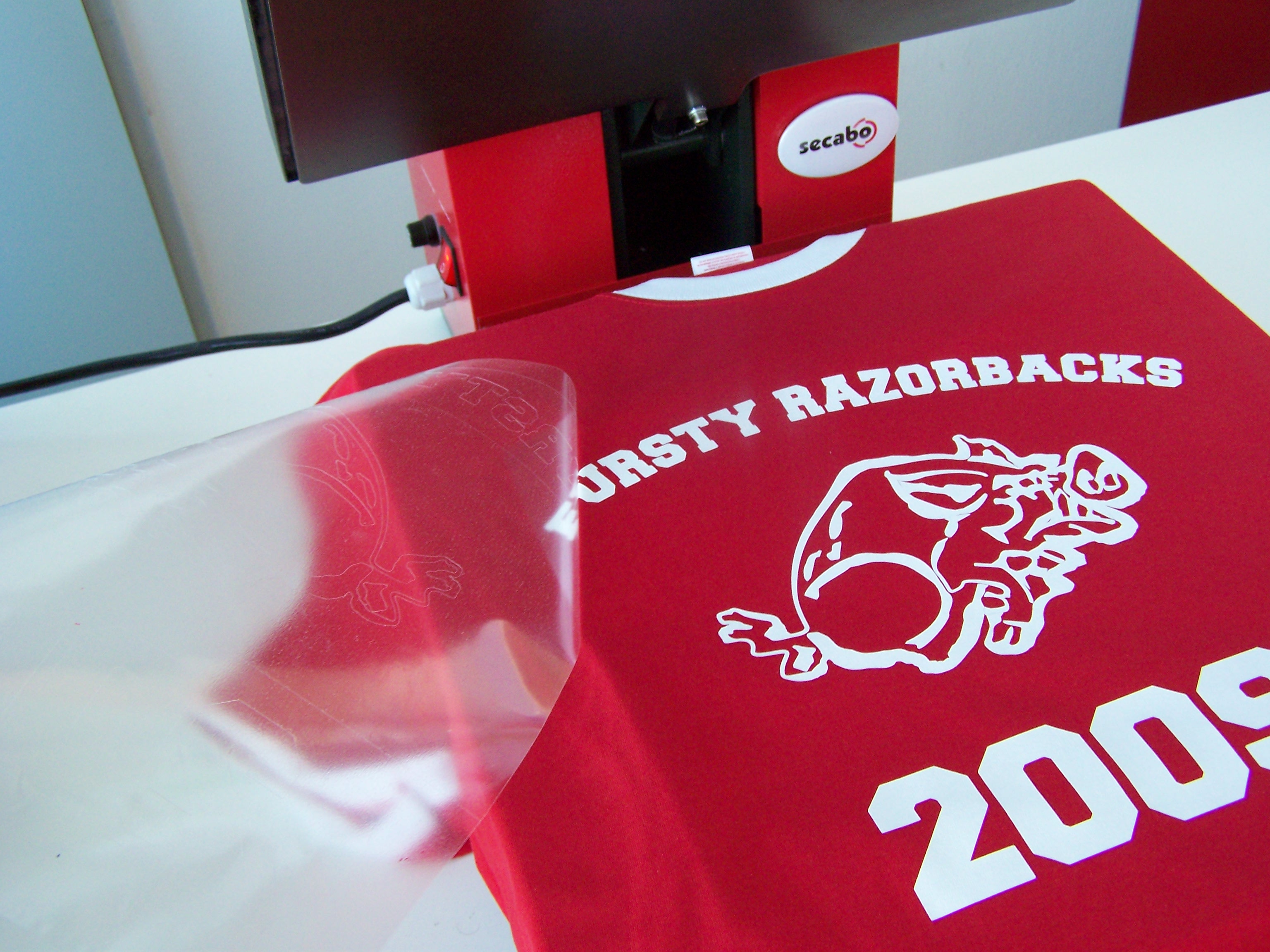 Transparente Trägerfolie wird von rotem T-Shirt mit weißem Flexfolien-Motiv Razorbacks 2009 abgezogen, dahinter Secabo-Transferpresse
