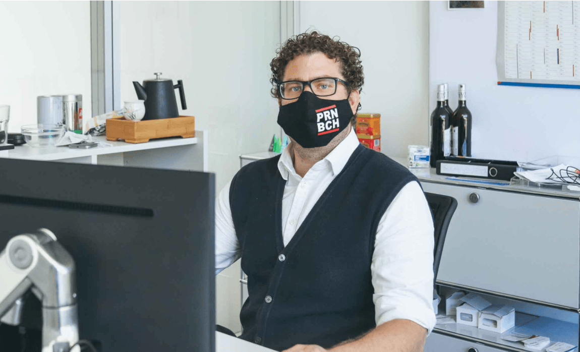 A man with curly hair and glasses is wearing a black cloth face mask with ‘PRN BCH’ printed on it and is sitting at a desk in front of a monitor in an office