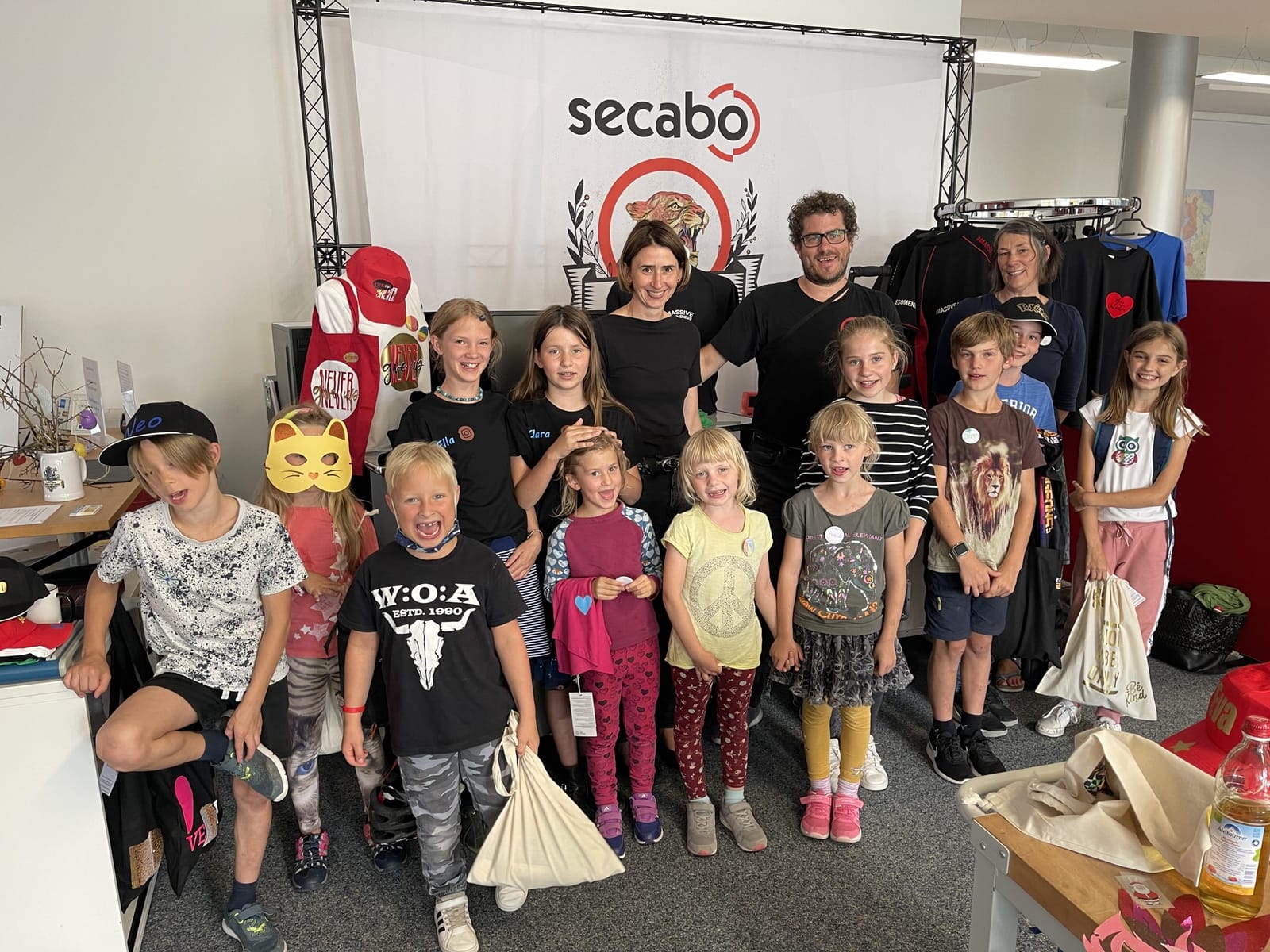 Children from Pörnbach taking part in the ‘Ferienpass’ programme, with three supervisors, posing in front of a Secabo banner, wearing T-shirts, caps, badges and cloth bags they had designed themselves