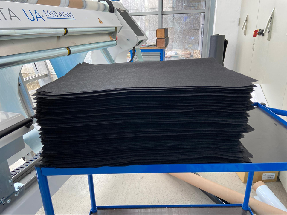 Pile de découpes de non-tissé noir coupées à longueur, posées sur un chariot bleu devant le centre de transformation NEPATA UA1650 ADWS